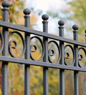 Black wrought iron fence with decorative scrollwork and finials in front of autumn trees